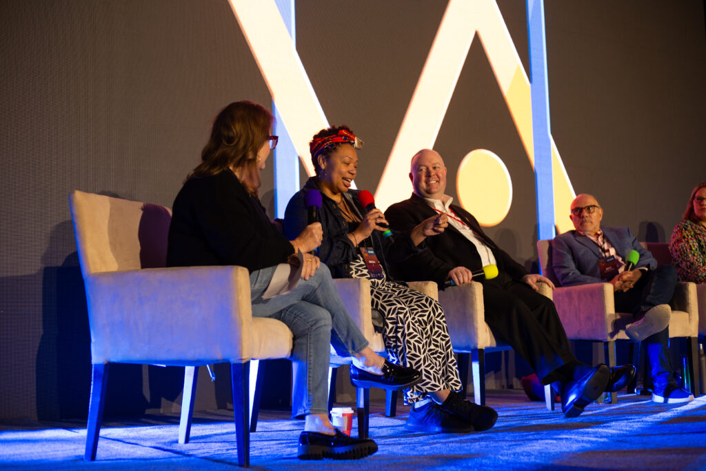GaRRS Goes to the VO Atlanta Conference 2025 2 A Panel Of Voice-Over Professionals Speak And Laugh During A Main Stage Discussion At Vo Atlanta 2025. One Woman Speaks Into A Red Microphone, Surrounded By Fellow Panelists Holding Microphones In Various Colors. The Large Vo Atlanta Logo Is Visible On The Screen Behind Them.
